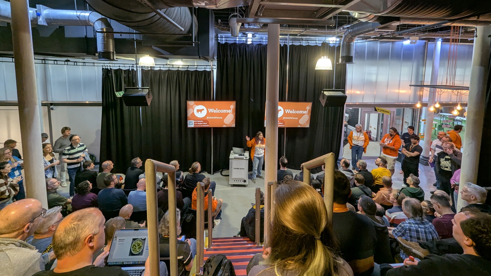A large group of attendees gather for the opening keynote at the DDD South West conference. The room has industrial-style ceilings with large vents and pipes, and two screens display a 'Welcome!' message. Some staff members in orange jackets stand near the stage while the audience listens from rows of tiered seating. Click to open full size image.