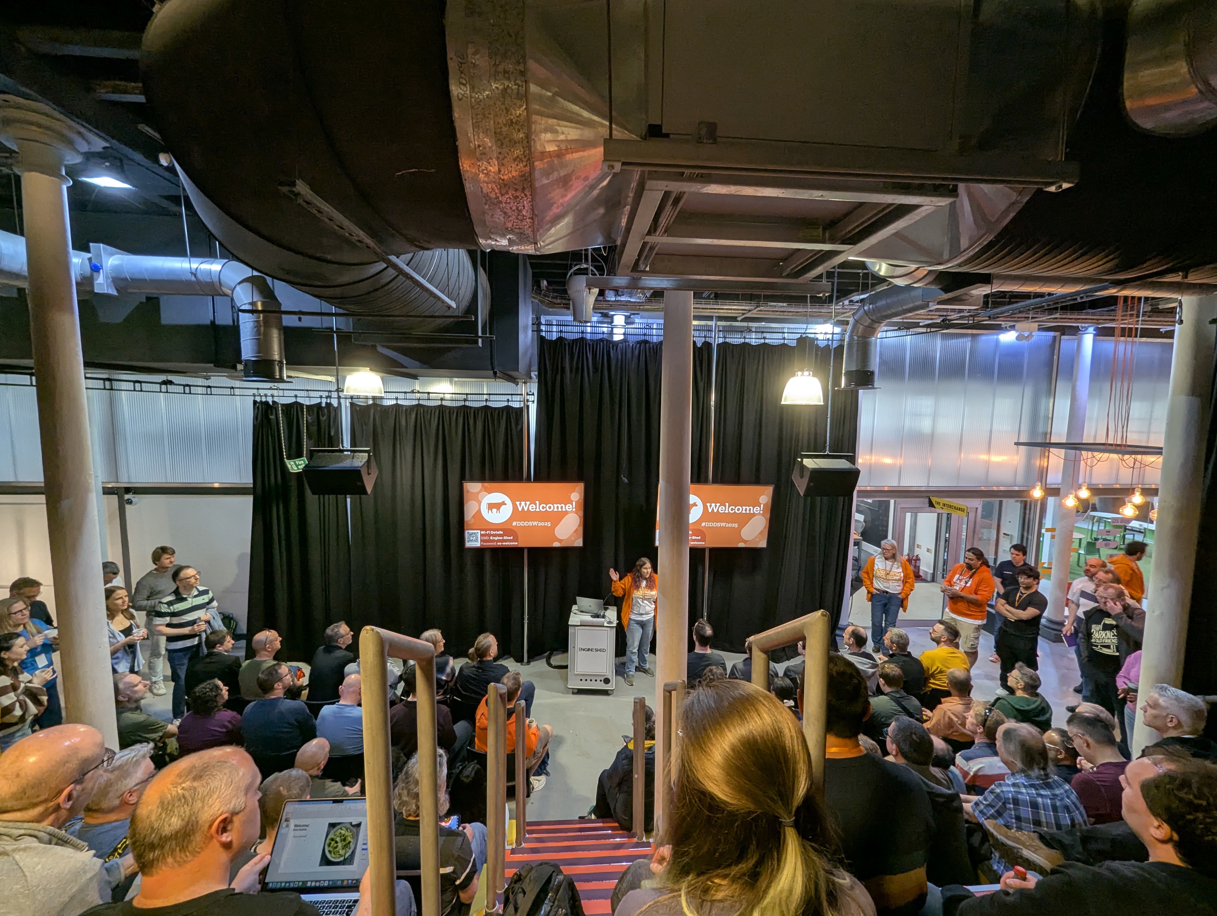 A large group of attendees gather for the opening keynote at the DDD South West conference. The room has industrial-style ceilings with large vents and pipes, and two screens display a 'Welcome!' message. Some staff members in orange jackets stand near the stage while the audience listens from rows of tiered seating.