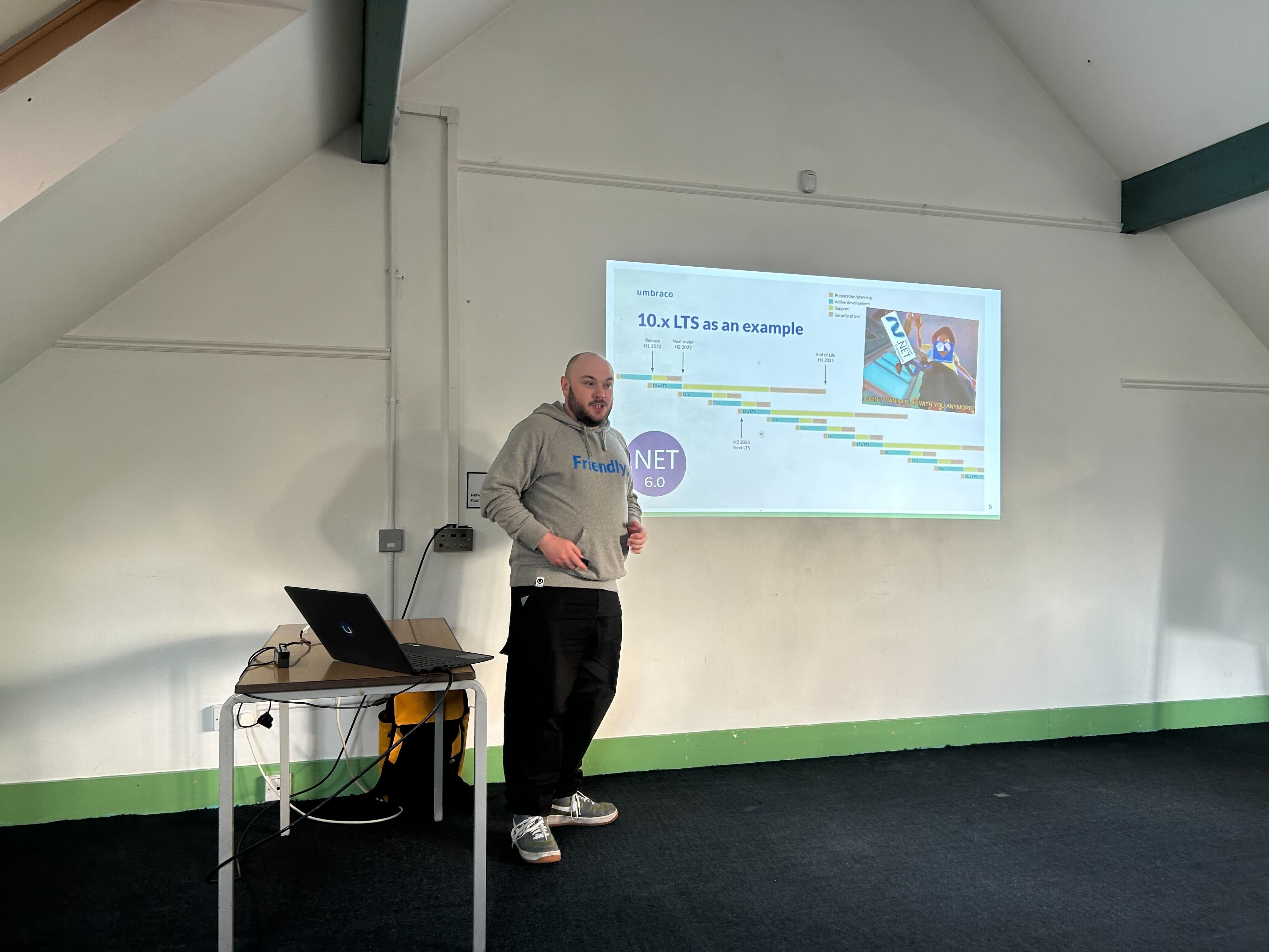 A speaker (Owain) stands in front of a projected slide at the umBristol April Umbraco meetup, smiling while presenting. He wears a grey 'Friendly' hoodie and dark trousers. A laptop sits on a small table nearby, connected to the projector. The room has a sloped ceiling and green trim along the walls.
