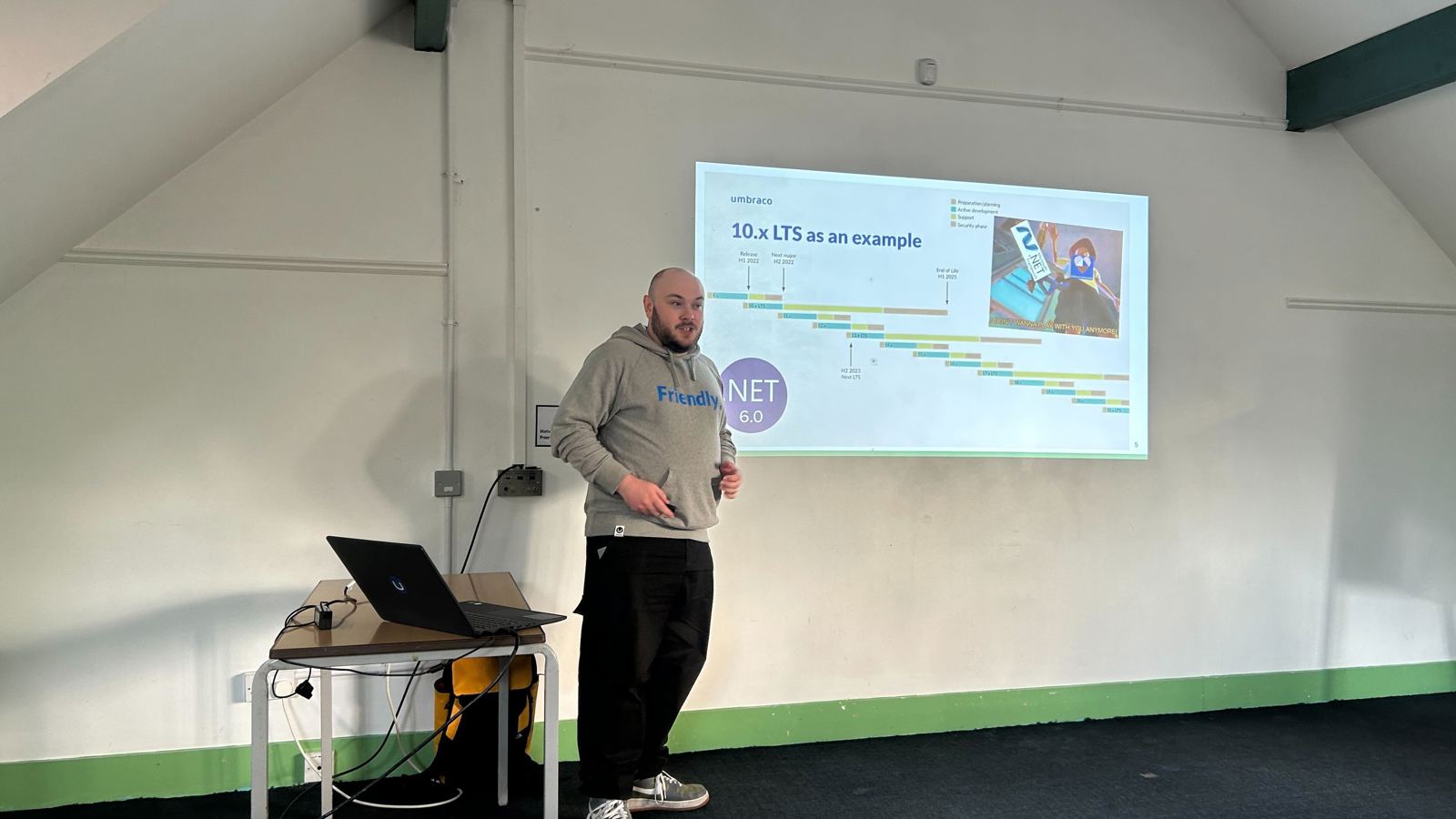 A speaker (Owain) stands in front of a projected slide at the umBristol April Umbraco meetup, smiling while presenting. He wears a grey 'Friendly' hoodie and dark trousers. A laptop sits on a small table nearby, connected to the projector. The room has a sloped ceiling and green trim along the walls. Click to open full size image.