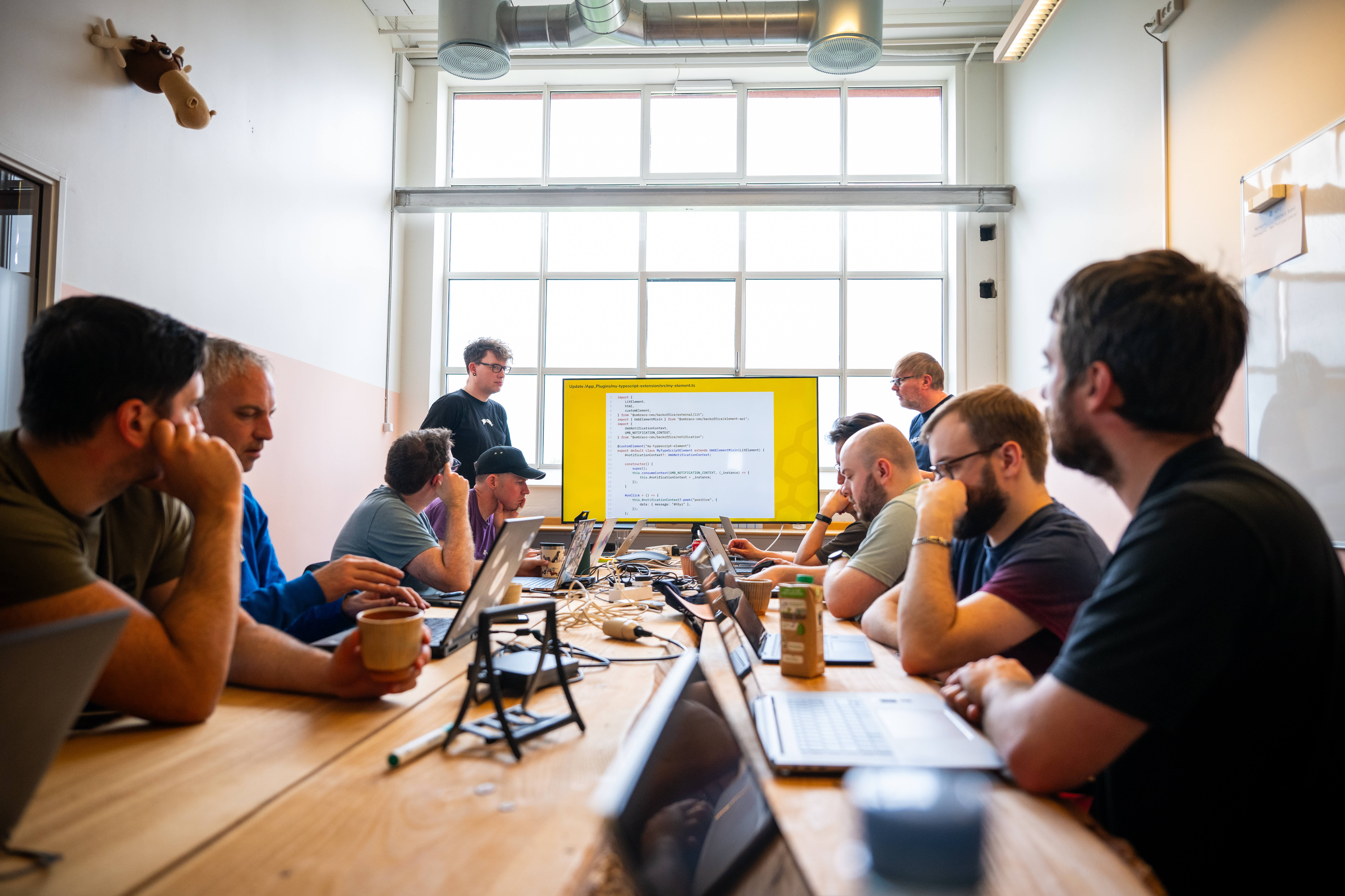 A group of people are sitting around a long wooden table in a well-lit room with a large window in the background. They are engaged in a meeting or workshop, with laptops and notebooks in front of them. Two individuals are standing at the front of the room, presenting a slide on a large screen that displays code. The attendees appear focused and attentive, suggesting a technical or coding workshop. The room has a modern industrial look with exposed ducts on the ceiling. Click to open full size image.