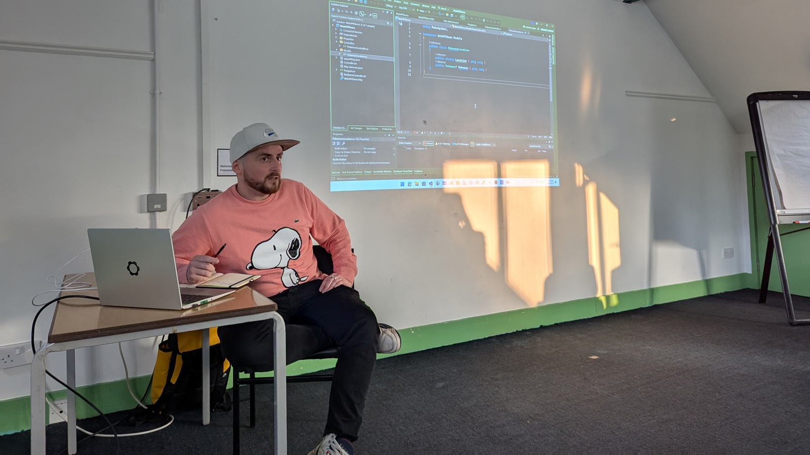 A speaker (Luke) sits in front of a projected slide at the umBristol April Umbraco meetup, smiling while presenting. He wears a pink snoopy hoodie and dark trousers. A laptop sits on a small table nearby, connected to the projector. The room has a sloped ceiling and green trim along the walls. Click to open full size image.