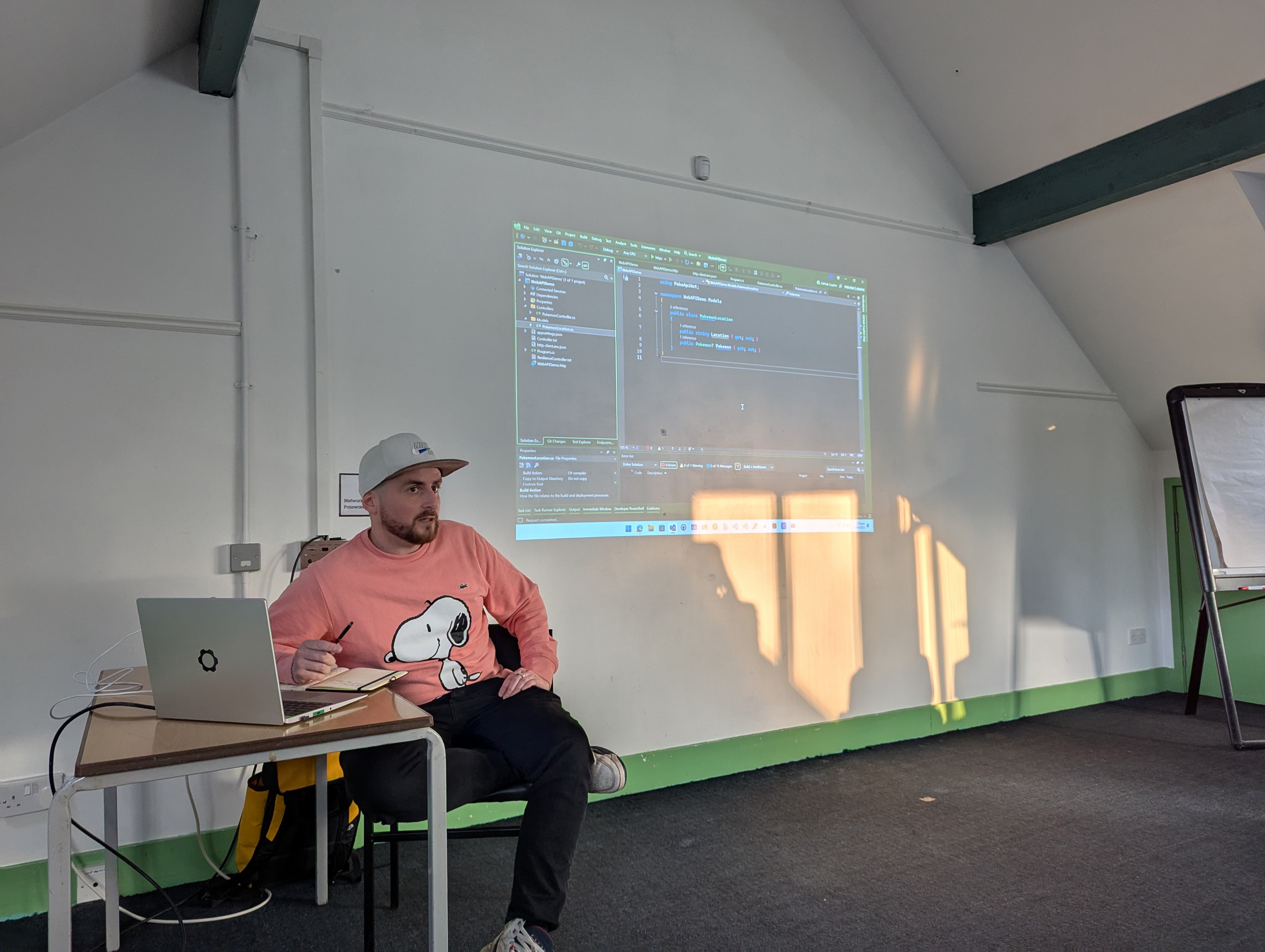 A speaker (Luke) sits in front of a projected slide at the umBristol April Umbraco meetup, smiling while presenting. He wears a pink snoopy hoodie and dark trousers. A laptop sits on a small table nearby, connected to the projector. The room has a sloped ceiling and green trim along the walls.