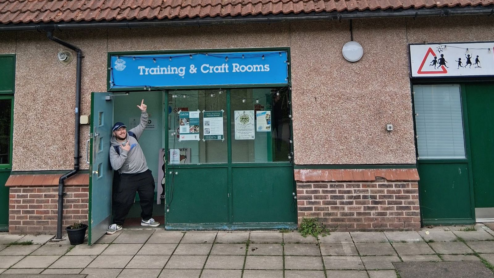 Owain standing in the doorway of a building, pointing and smiling at a sign that says "Training & Craft rooms" Click to open full size image.