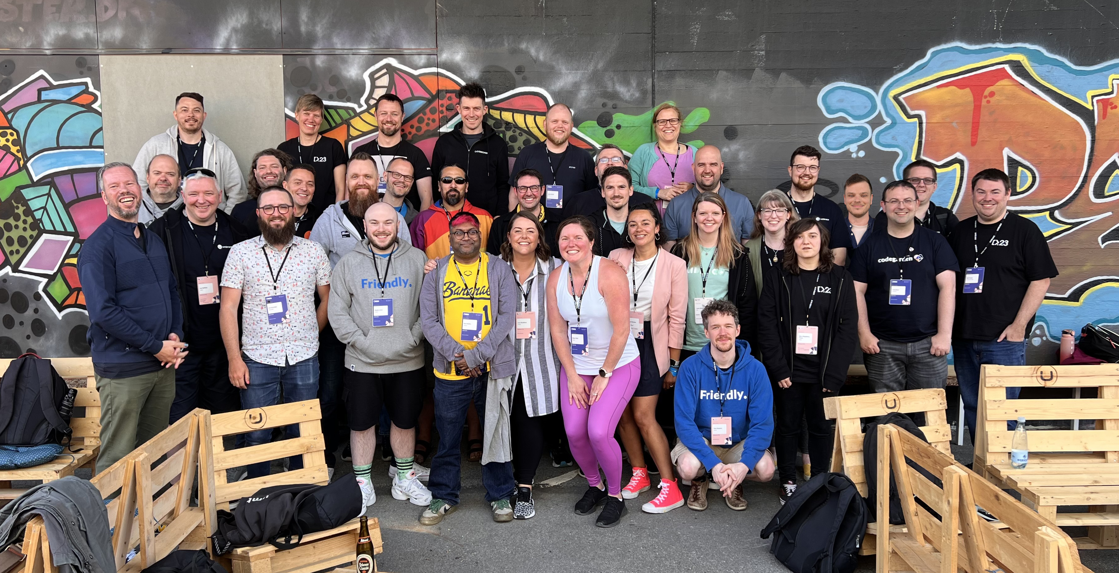 A group photo of CODECABIN attendees taken at the Codegarden conference. The group in the phot consists of around 33 people, standing and seated in an outdoor setting against a wall adorned with colorful graffiti art. The attendees are smiling and appear to be in high spirits, many wearing conference lanyards and badges. They are casually dressed, with some wearing branded t-shirts and hoodies. The seating area is made up of wooden pallet benches. The atmosphere is vibrant and energetic.