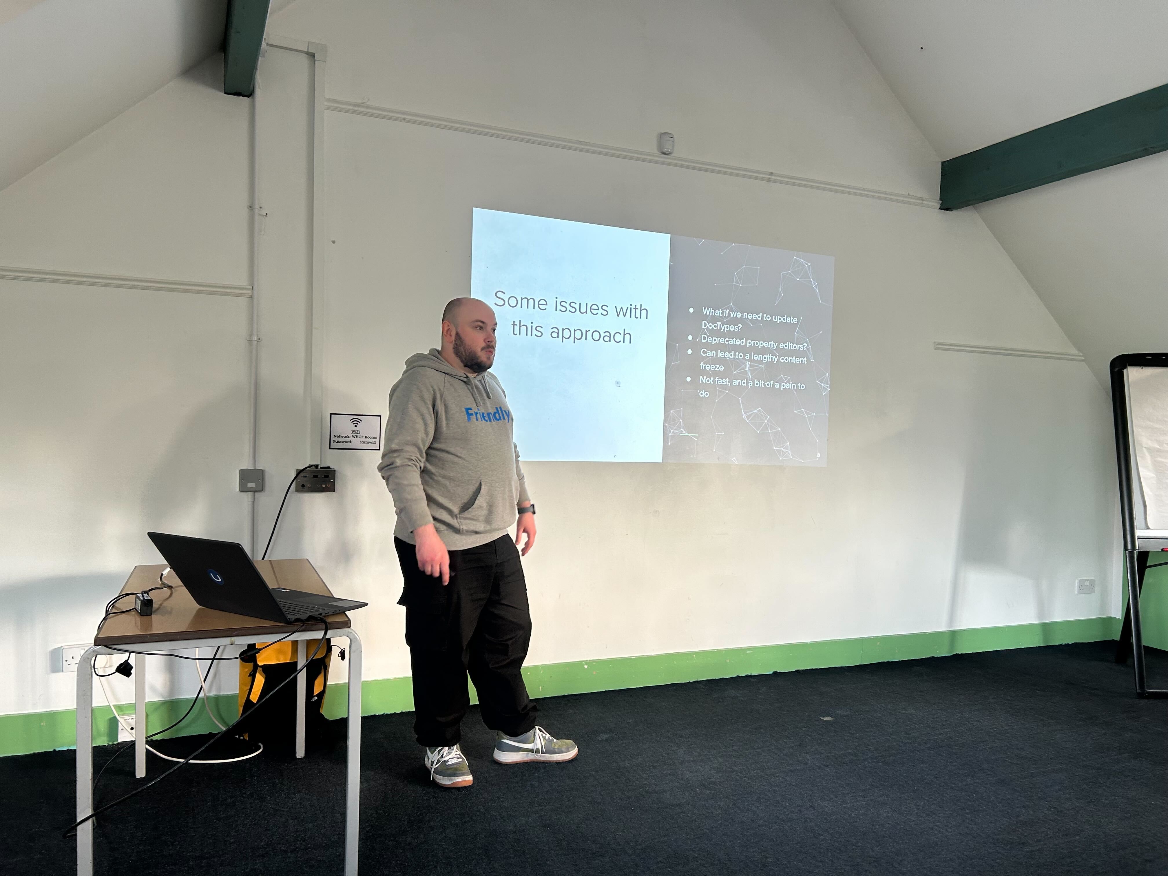 A speaker (Owain) stands in front of a projected slide at the umBristol April Umbraco meetup, smiling while presenting. He wears a grey 'Friendly' hoodie and dark trousers. A laptop sits on a small table nearby, connected to the projector. The room has a sloped ceiling and green trim along the walls.