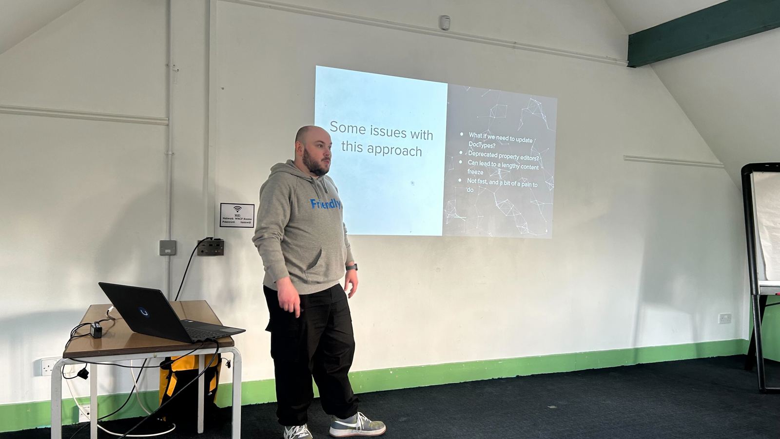 A speaker (Owain) stands in front of a projected slide at the umBristol April Umbraco meetup, smiling while presenting. He wears a grey 'Friendly' hoodie and dark trousers. A laptop sits on a small table nearby, connected to the projector. The room has a sloped ceiling and green trim along the walls. Click to open full size image.
