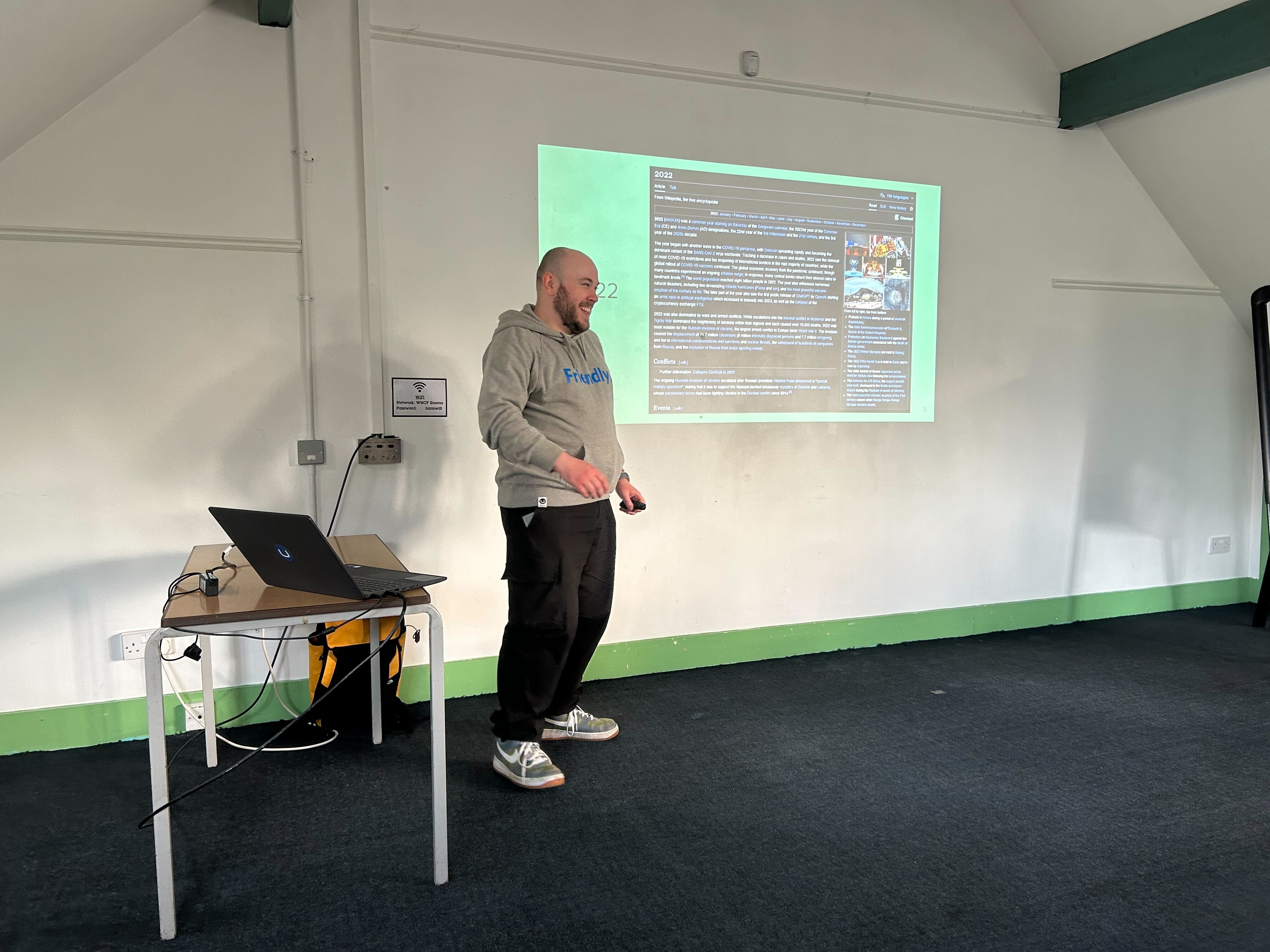A speaker (Owain) stands in front of a projected slide at the umBristol April Umbraco meetup, smiling while presenting. He wears a grey 'Friendly' hoodie and dark trousers. A laptop sits on a small table nearby, connected to the projector. The room has a sloped ceiling and green trim along the walls.