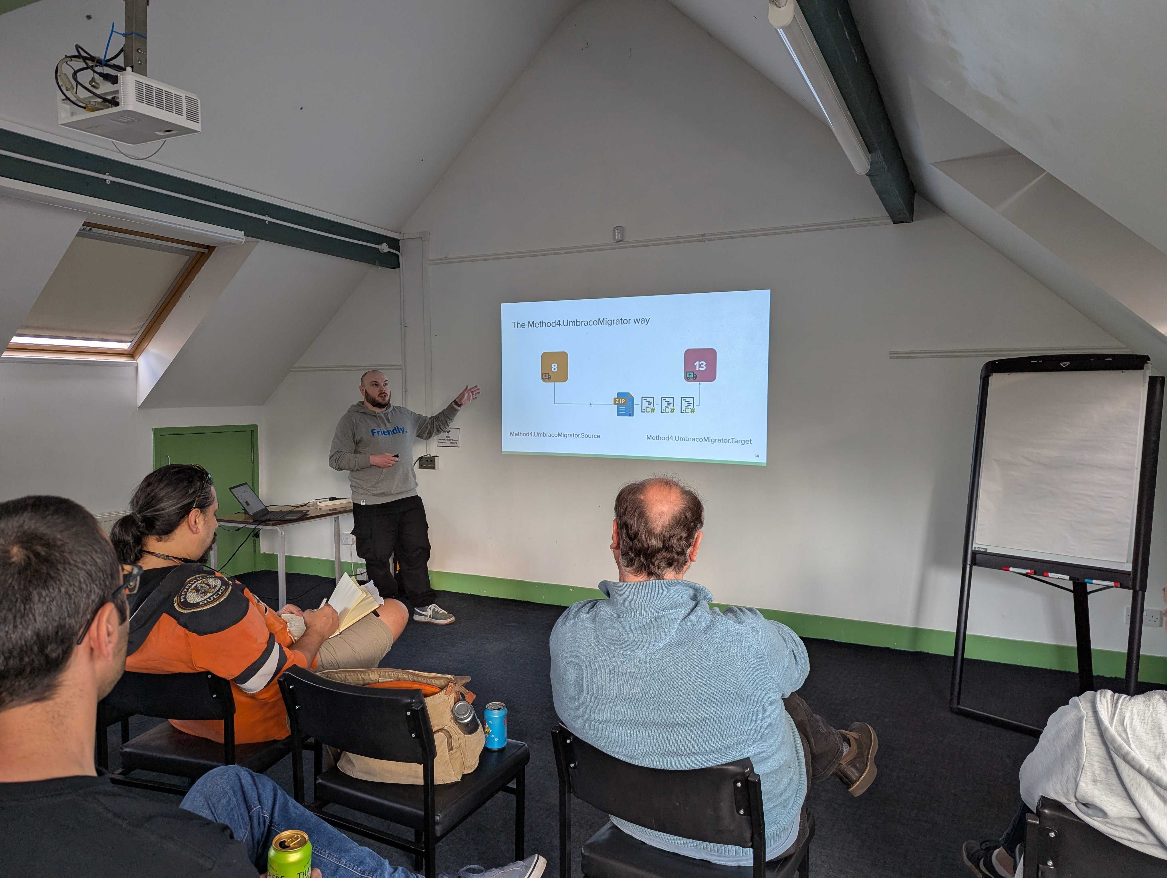 A speaker gives a presentation at the umBristol April Umbraco meetup, standing next to a projected slide titled 'The Method4UmbracoMigrator way.' Several attendees sit facing the presenter, who gestures toward the slide. The room has a sloped ceiling, a skylight, and a blank flip chart nearby.