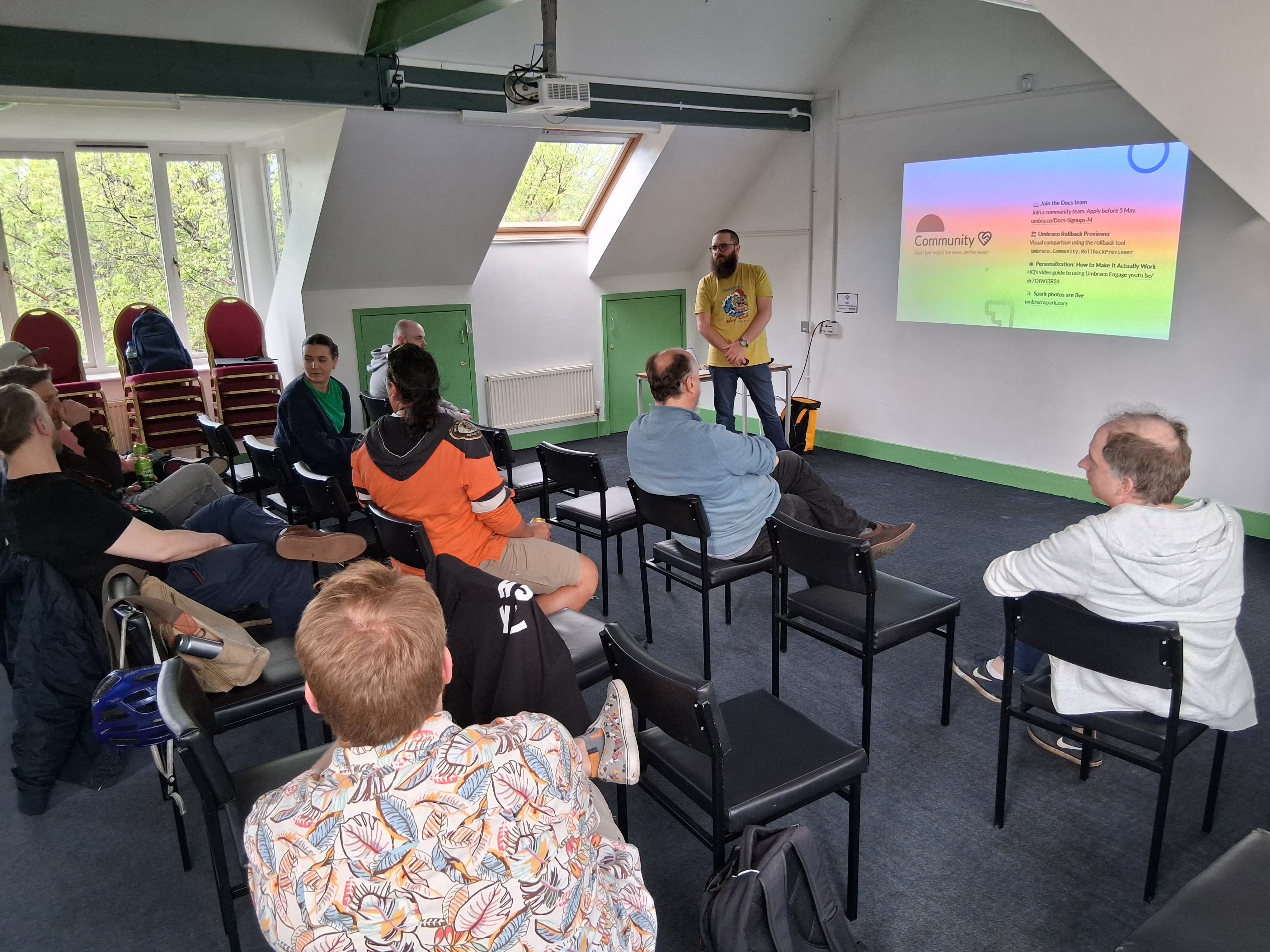 A speaker (Joe) stands in front of a projected slide at the umBristol April Umbraco meetup, smiling while presenting. He wears a yellow shirt and dark blue jeans. A laptop sits on a small table nearby, connected to the projector. The room has a sloped ceiling, windows with trees visible outside, and green trim along the walls.