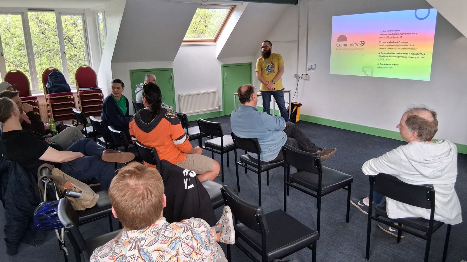 A speaker (Joe) stands in front of a projected slide at the umBristol April Umbraco meetup, smiling while presenting. He wears a yellow shirt and dark blue jeans. A laptop sits on a small table nearby, connected to the projector. The room has a sloped ceiling, windows with trees visible outside, and green trim along the walls. Click to open full size image.