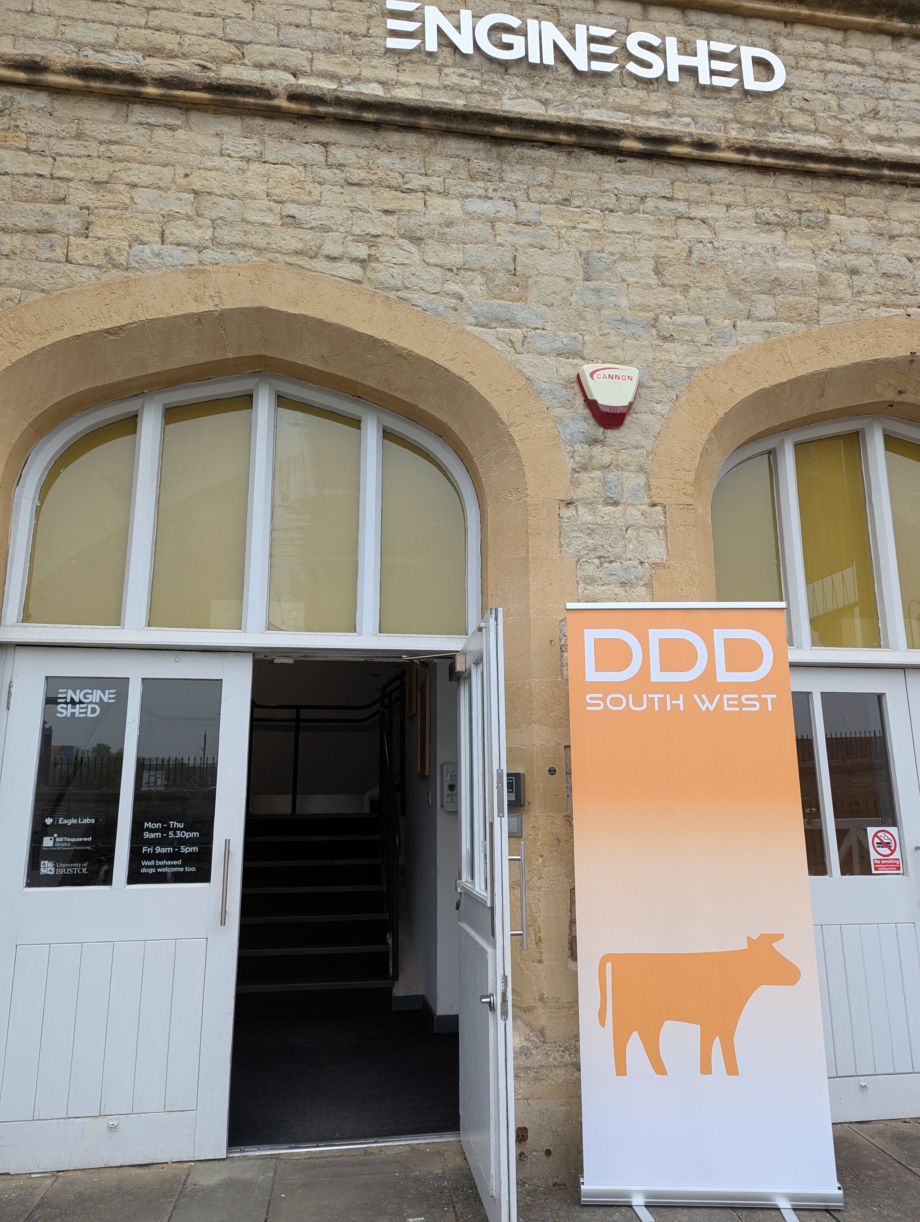 An image of the front door to the Engine Shed building, there is a popup sign by the side of the door that reads 'DDD South West' with an outline of a cow.' Click to open full size image.