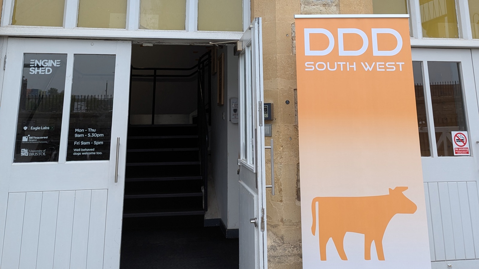 An image of the front door to the Engine Shed building, there is a popup sign by the side of the door that reads 'DDD South West' with an outline of a cow.' Click to open full size image.