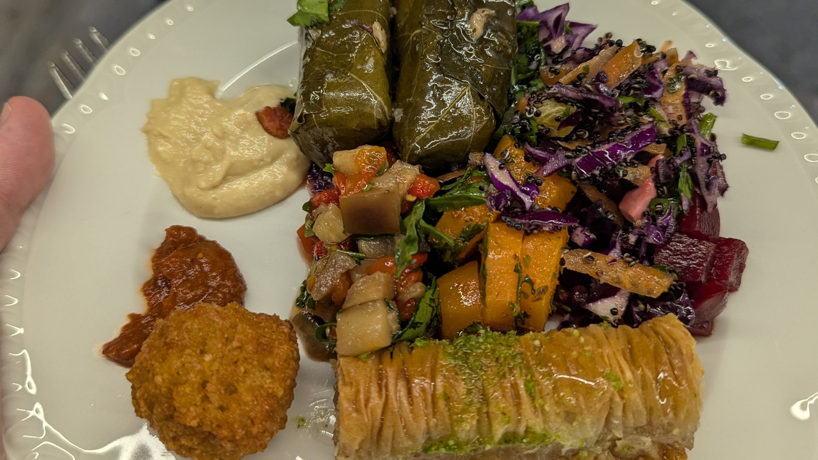 A close-up of a plate of Greek food at the DDD South West conference, featuring stuffed vine leaves, baklava, a colorful salad with vegetables, hummus, a small falafel ball, and a spicy red sauce. Click to open full size image.