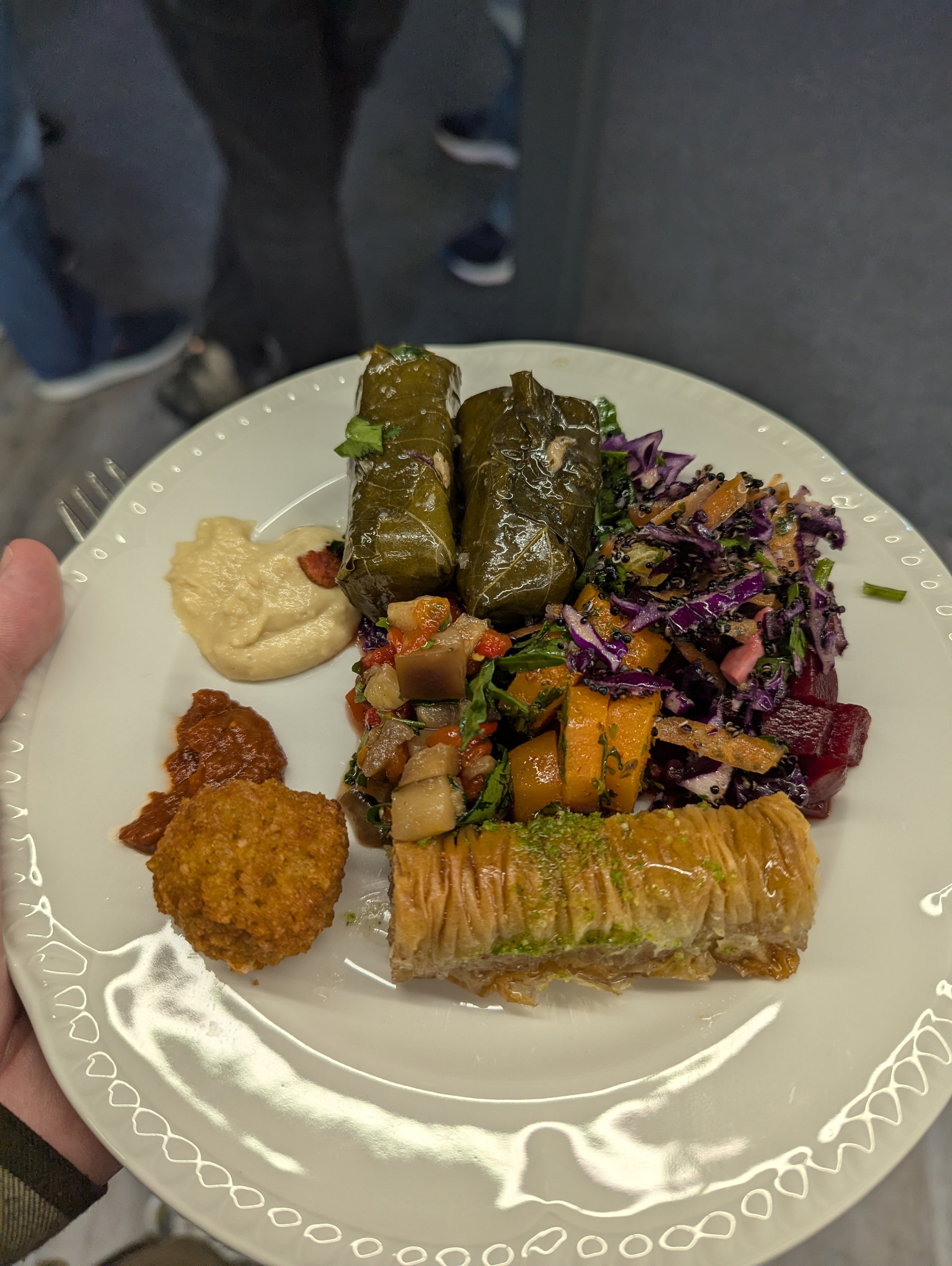 A close-up of a plate of Greek food at the DDD South West conference, featuring stuffed vine leaves, baklava, a colorful salad with vegetables, hummus, a small falafel ball, and a spicy red sauce.